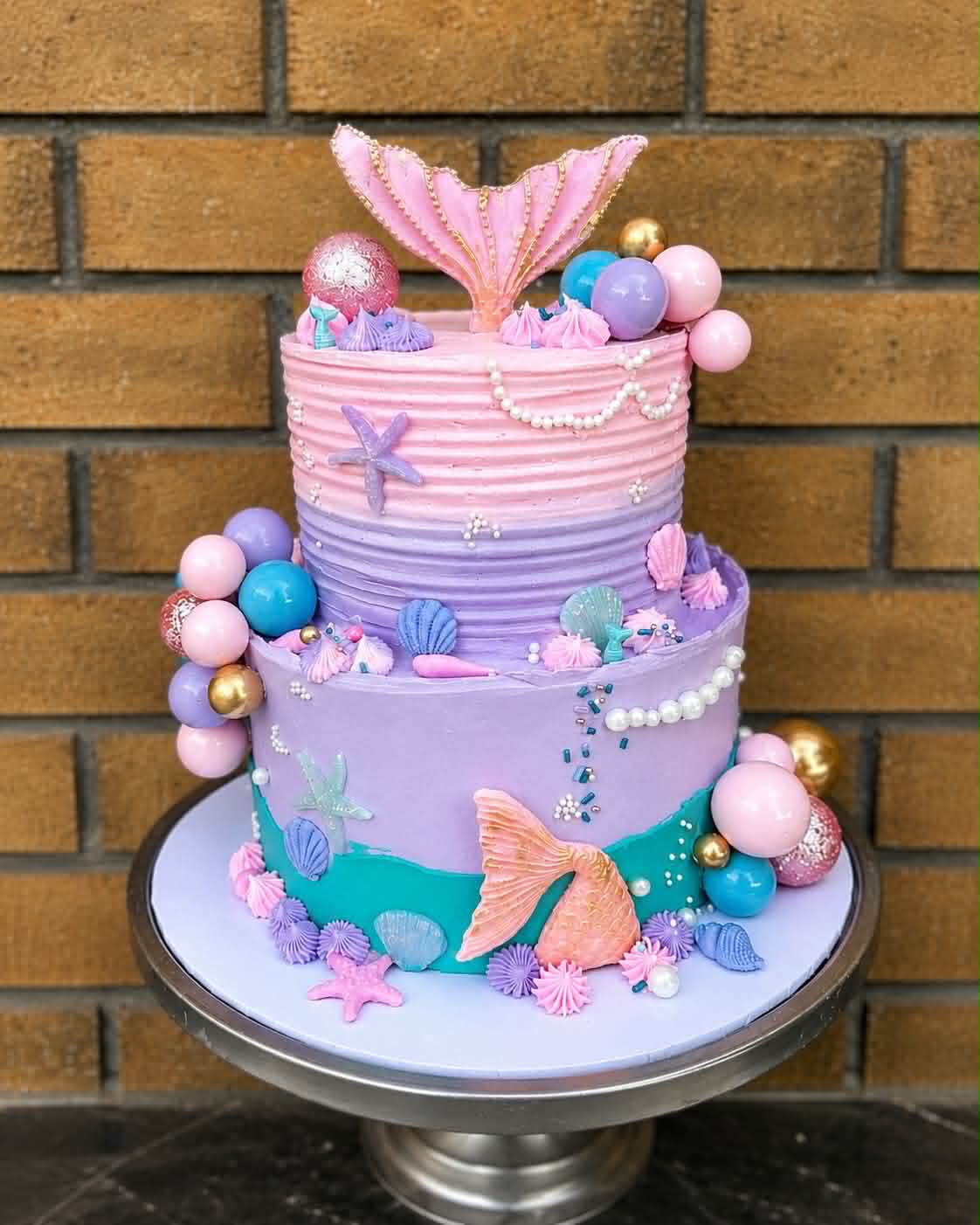 Two-tier mermaid birthday cake in pink, purple and teal with mermaid tail topper and seashell decorations